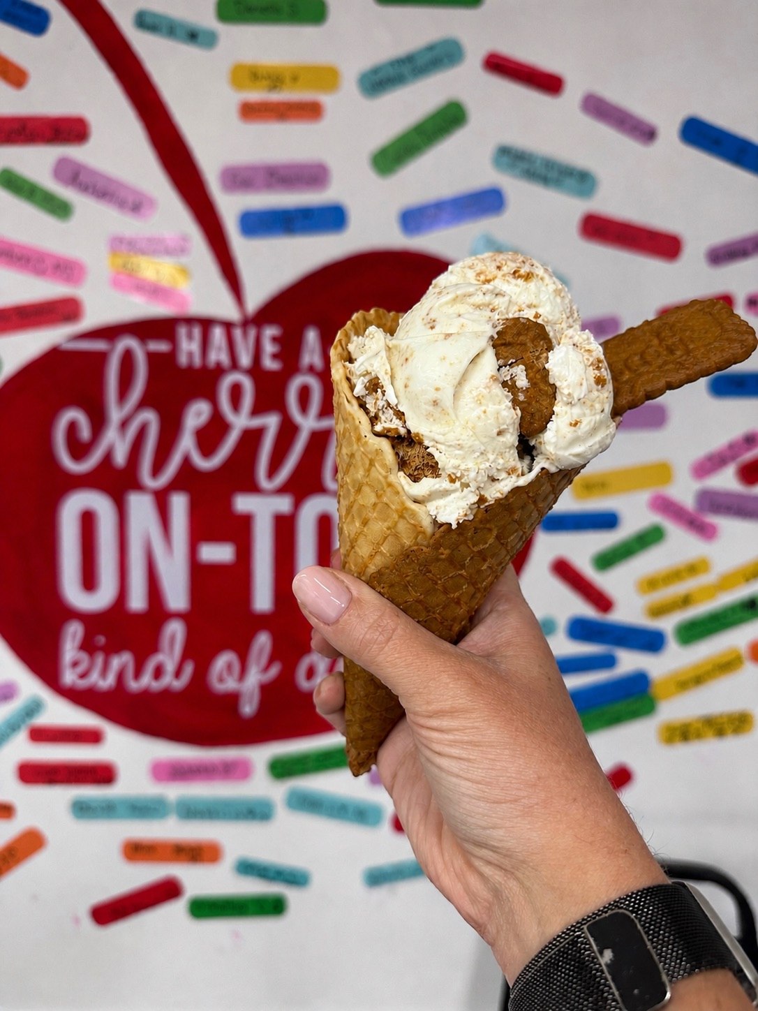 A hand-dipped scoop on a waffle cone held up in front of the shoppe's 'have a cherry on-top kind of day' sprinkle mural
