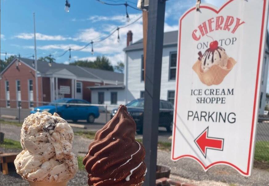 Two cones — one hand-dipped scoop, one chocolate-dipped soft serve — held up in front of the Cherry On Top parking sign